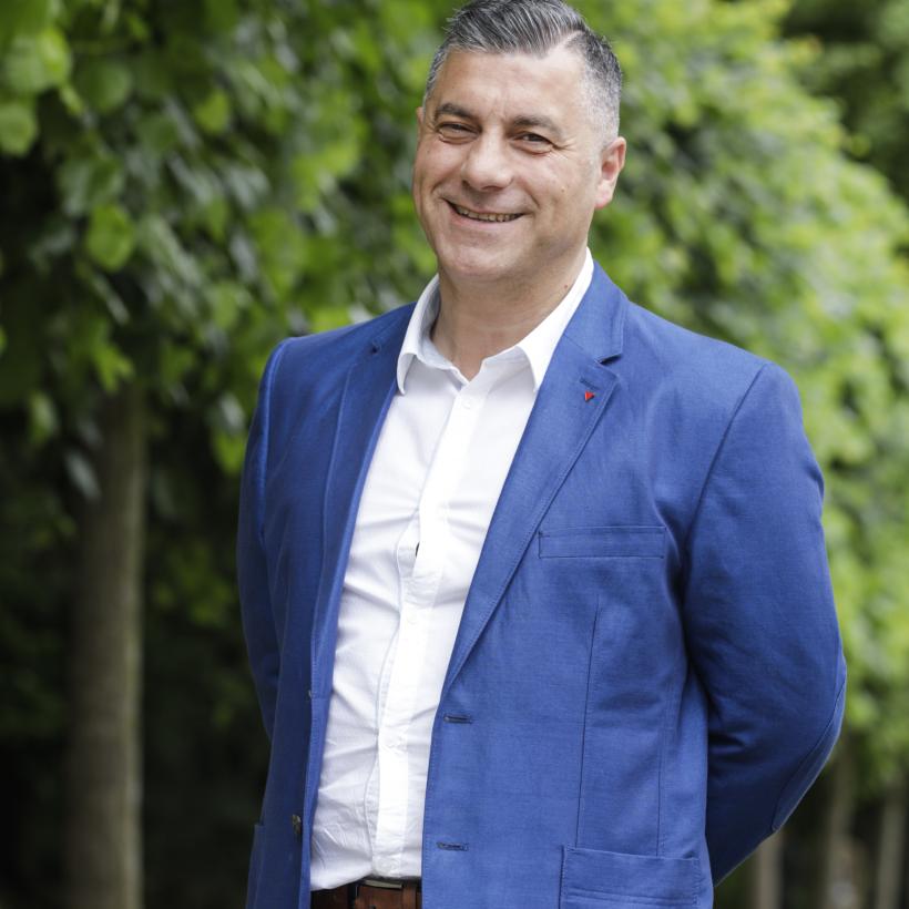 Roberto D'Amico se tient devant une rangée d'arbres. Il porte un blazer bleu foncé et une chemise blanche. Il a les mains derrière le dos. Il sourit amicalement.