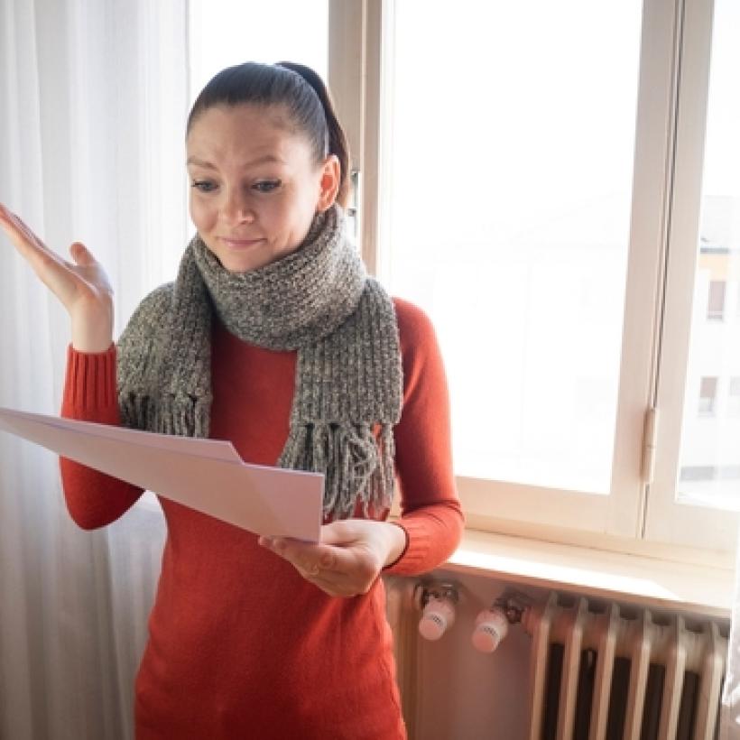 Een vrouw kijkt bezorgd naar een rekening. Ze draagt een grijze sjaal en een rood truitje. Ze staat met haar rug tegen de verwarming.