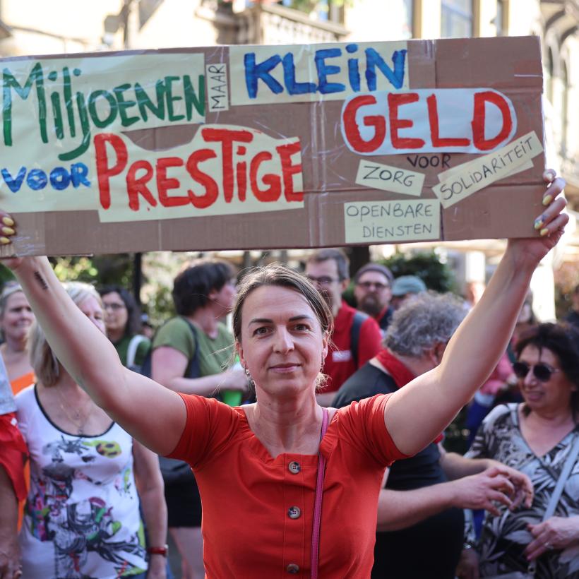 Vrouw protesteert tegen de besparingsplannen in Gent met een bord: “Miljoenen voor prestige, maar lein geld voor zorg, openbare diensten, solidariteit”