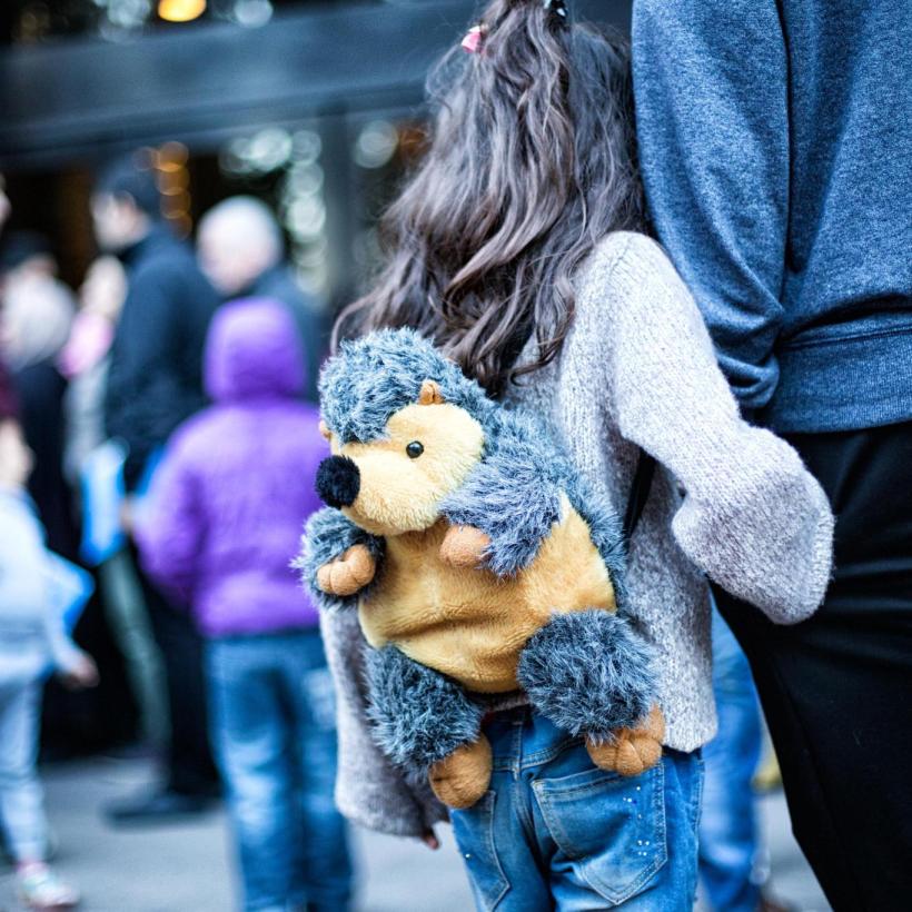 Een klein meisje, van achteren gezien, met een rugzak in de vorm van een egel en aan de hand van een volwassene