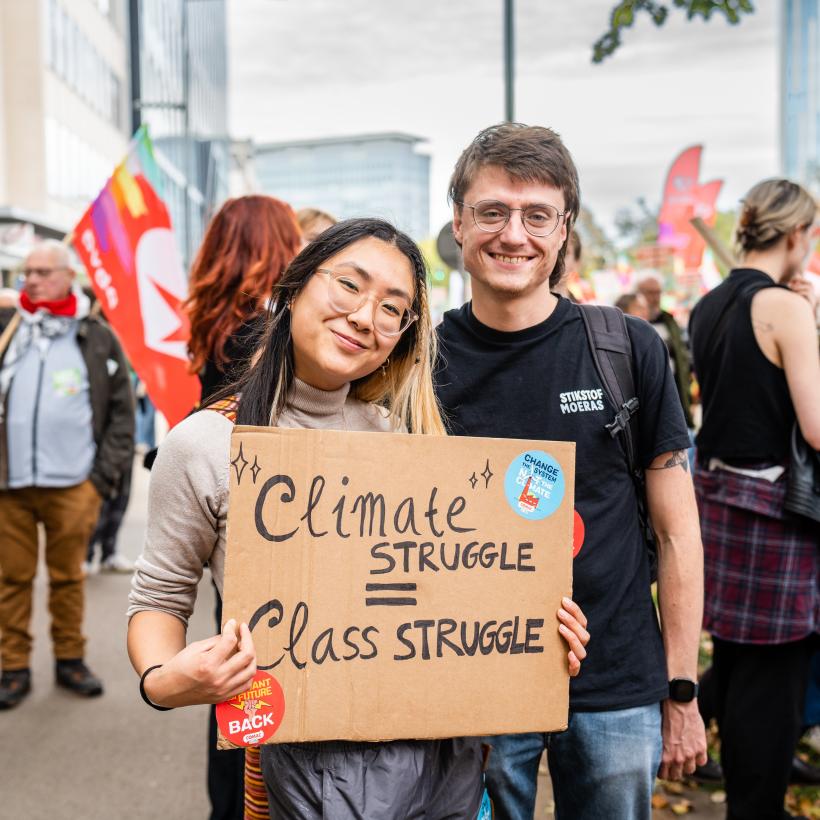 Een jonge man en een jonge vrouw bij een demonstratie houden een bordje vast met de tekst “Climate struggle = class struggle”