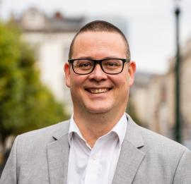 Raoul Hedebouw loopt op straat naast het Warandepark. Raoul lacht en kijkt richting de camera. Hij draagt een grijze blazer en een wit hemd.