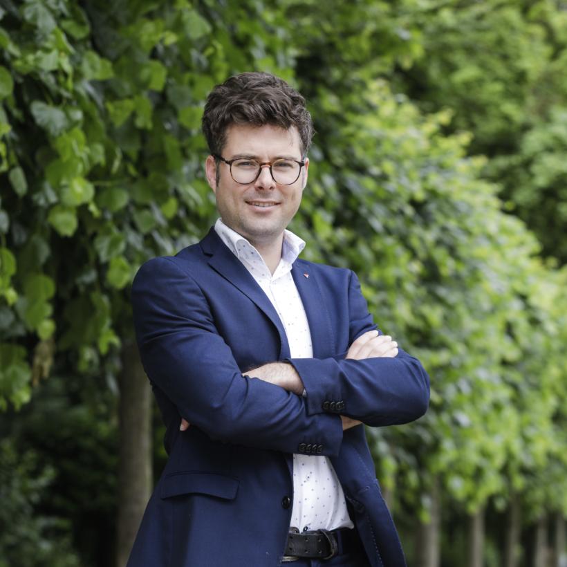 Steven De Vuyst se tient devant une rangée d'arbres. Il porte un blazer bleu foncé et une chemise blanche. Il sourit chaleureusement et a les bras croisés.