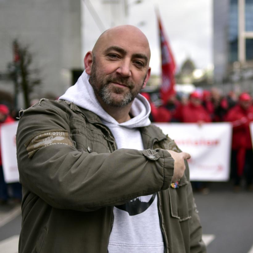 Gaby Colebunders kijkt lachend in de camera. Hij wijst achter zich naar vakbondsmanifestanten. Hij draagt een bruine jas en een lichtgrijze hoodie.