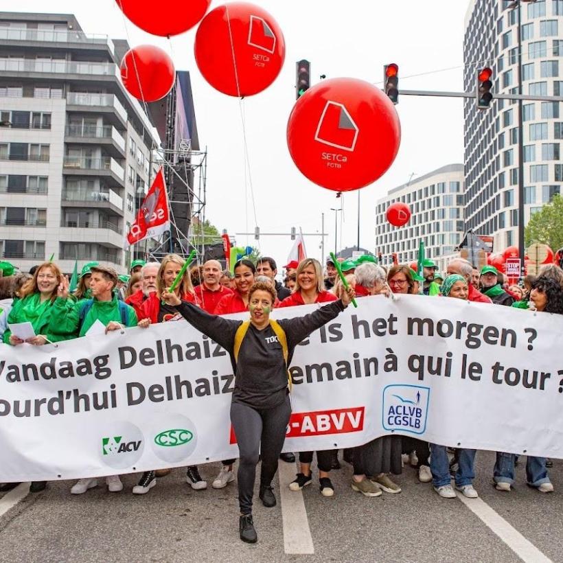 Vakbondsbetoging in Brussel. Een spandoek met daarop: "Vandaag Delhaize, aan wie is het morgen?" Voor het spandoek staat een persoon met de handen in de lucht en percussiestokken in de handen. Achter de spandoek staan vakbondsleden van het ACV en het ABVV