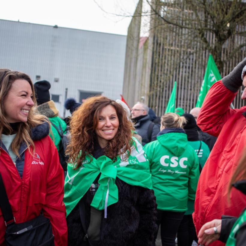 Deux travailleuses de Delhaize se trouvent au milieu d'une manifestation syndicale à Bruxelles. L'une porte une veste rouge FGTB, l'autre verte CSC. Elles sont très souriantes. Autour d'elles, il y a beaucoup de syndicalistes qui participent.