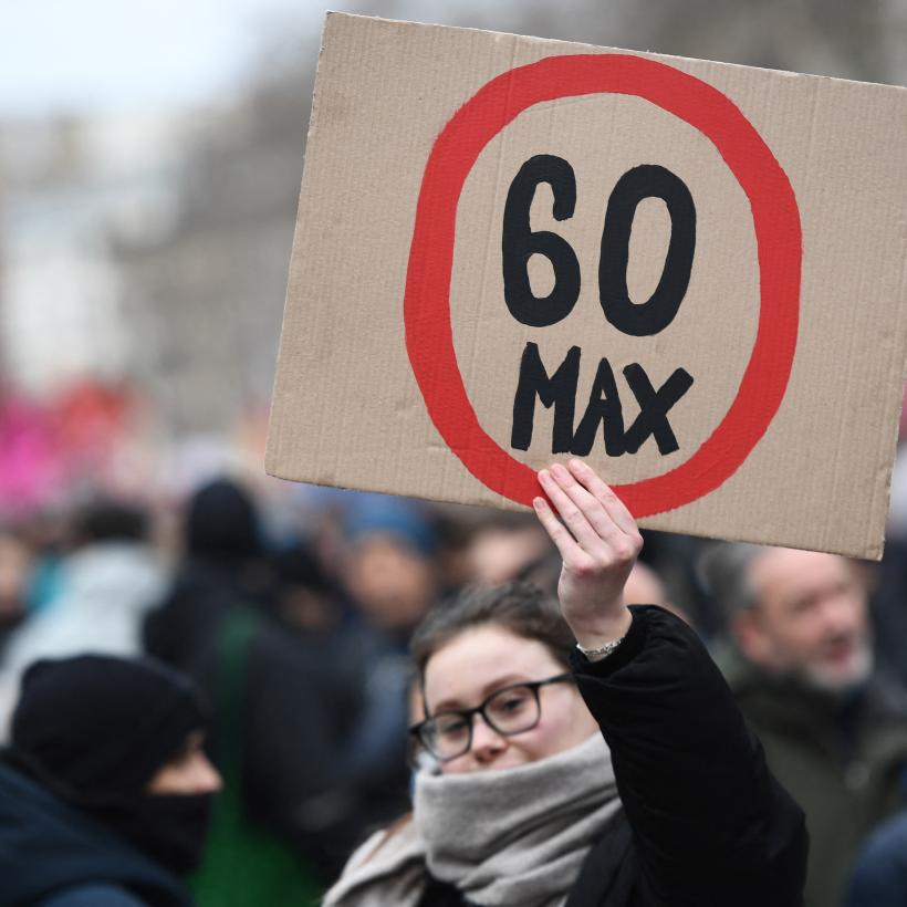 Vrouw op pensioenbetoging houdt kartonnen bord omhoog met verkeersbord '60 max' op geschilderd.