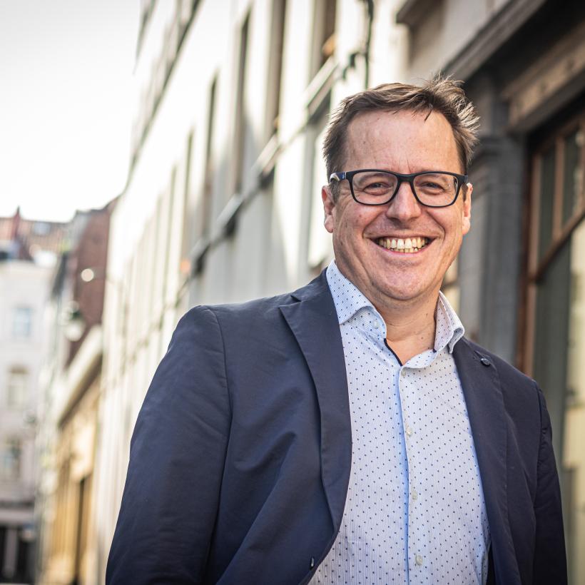 David Pestieau est photographié jusqu'à la moitié du torse. Il porte une chemise bleu clair et un blazer bleu foncé. Il sourit de toutes ses dents. Derrière lui, on aperçoit des maisons. 