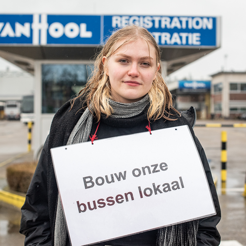 Een jonge vrouw staat voor de Van Hool fabriek met een bord waarop staat "Bouw onze bussen lokaal".
