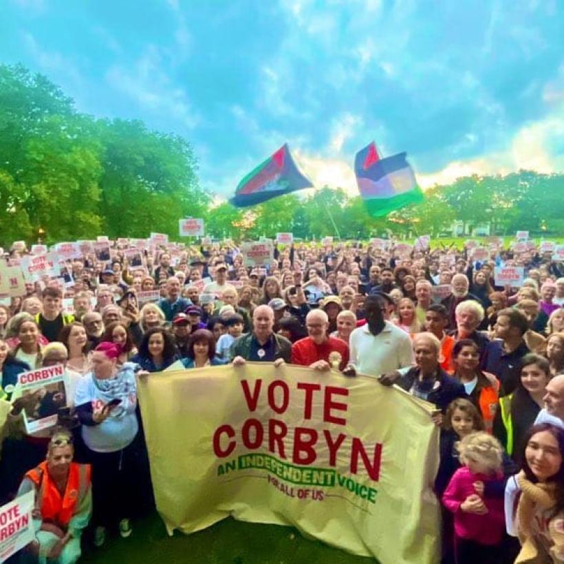 Jeremy Corbyn staat voor een menigte. Een vlag met "Vote Corbyn" hangt voor hen. 