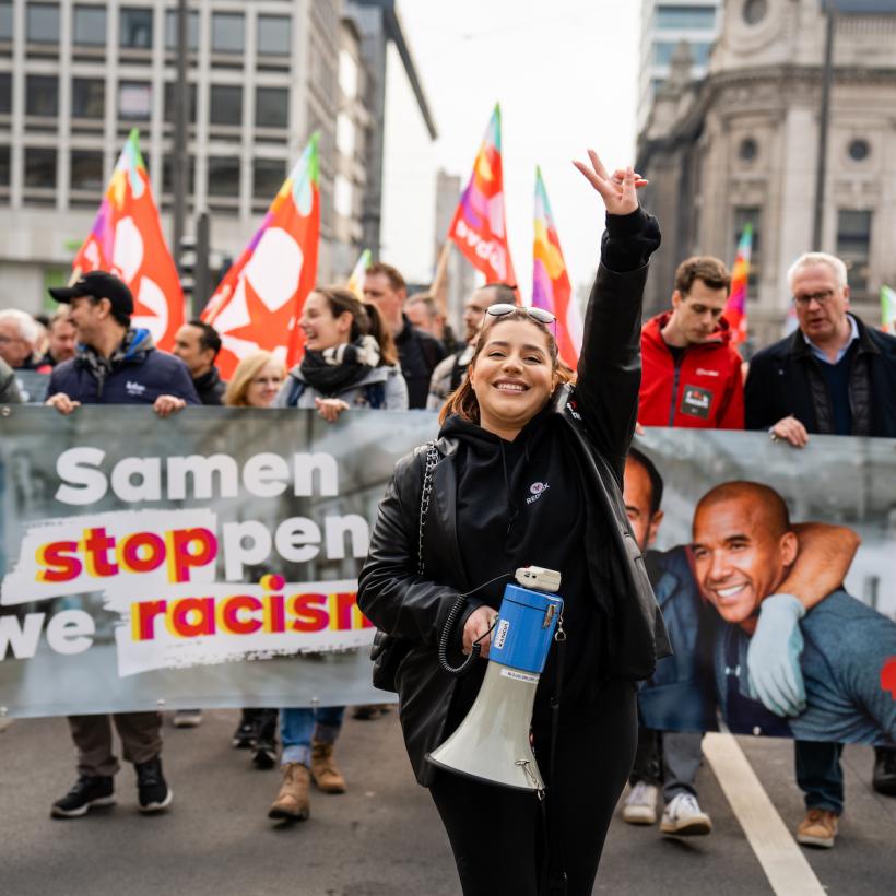 Een jonge vrouw tijdens een anitracismebetoging. Ze steekt twee vingers in de lucht. Achter haar een blok van de PVDA met een spandoek met daarop ‘Samen stoppen we racisme’