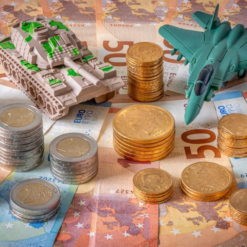 Er liggen eurobiljetten, stapels munten en een plastieken miniatuurtank en -gevechtsvliegtuig op tafel.